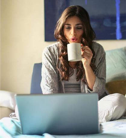 Lady sitting in bed on her computer shopping for mattresses. She is blowing into a hot drink, while looking down at her computer screen. 